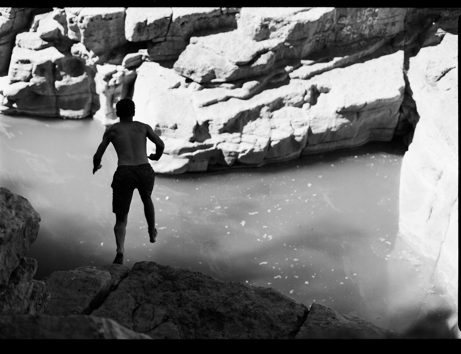 Black and white photo of a man jumping off a rocky cliff into a river in Morocco.